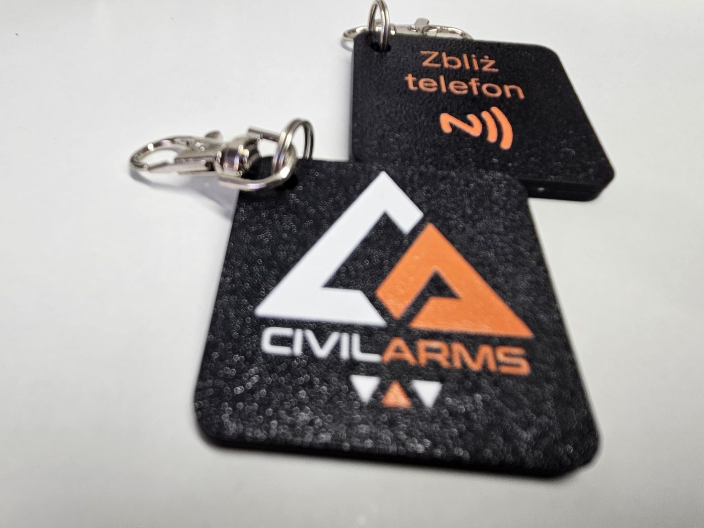 Czarny brelok z kolorowym logo Civil Arms oraz brelok z pomarańczowym napisem Zbliż telefon i ikoną NFC