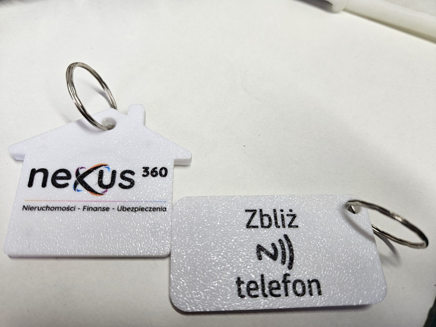 Brelok w kształcie domku z logo Nexus360 oraz brelok z napisem Zbliż telefon i ikoną NFC