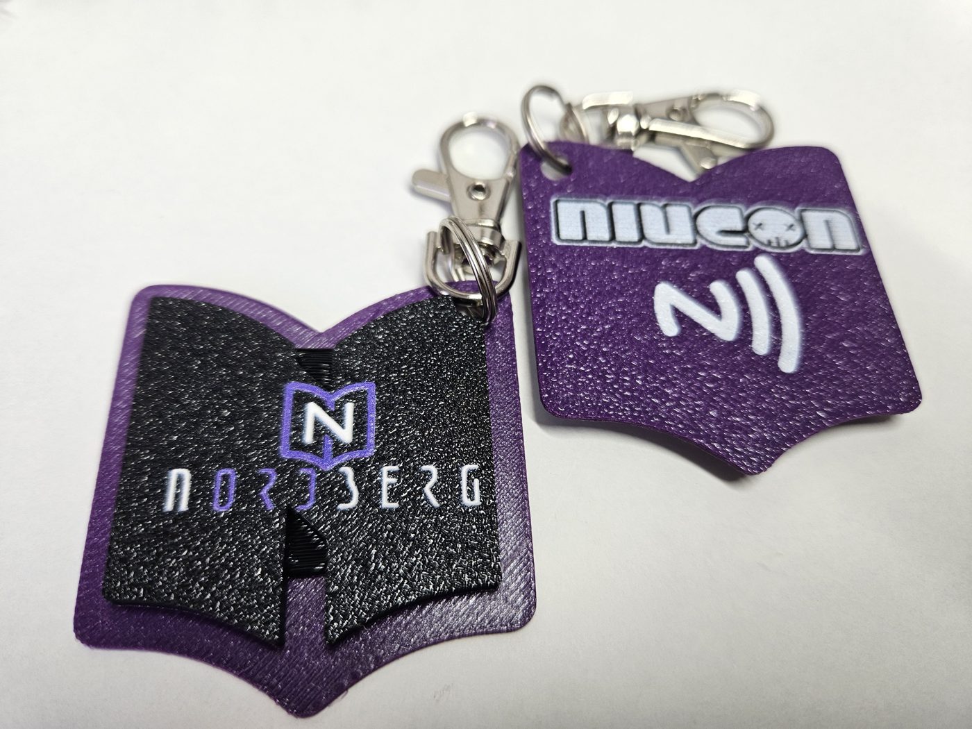 Dwa breloki NFC w kształcie otwartej książki z logo Nordberg i Niucon, fioletowo-czarne, z karabińczykami