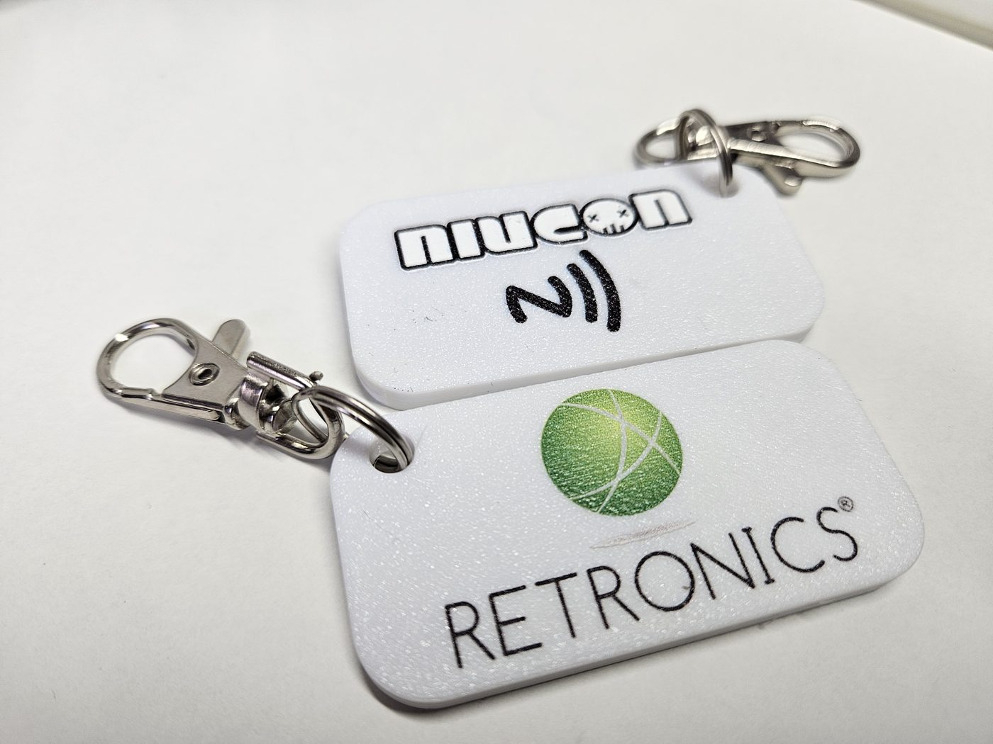 Dwa białe prostokątne breloki NFC z logo Niucon i Retronics oraz karabińczykami